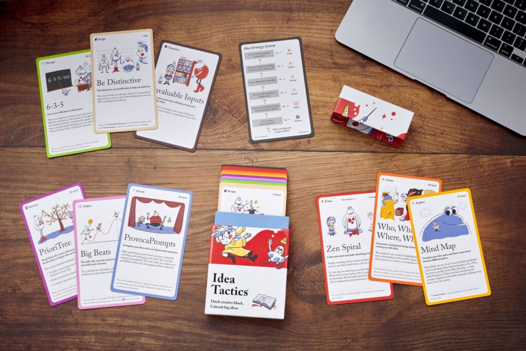 idea tactics box and cards spread out on a wooden table, with a Macbook in the corner. 